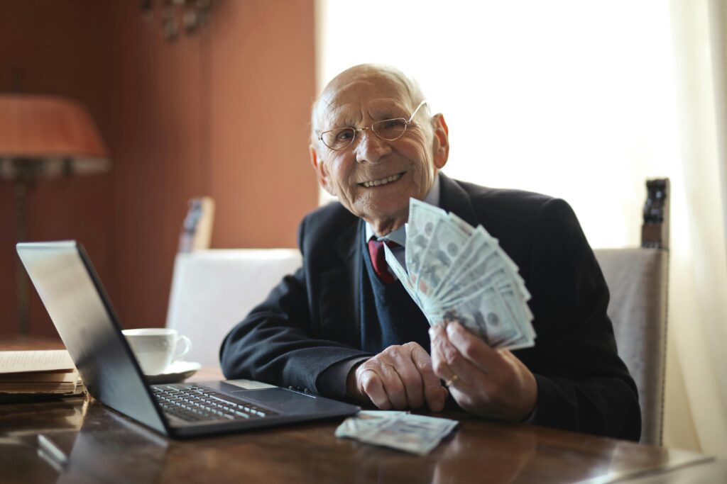 Passive income Happy senior businessman showing cash at his desk with a laptop, indoors.