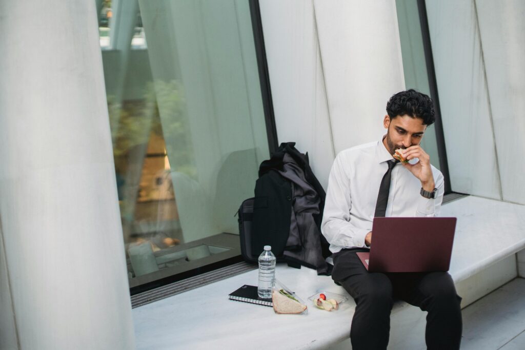 Businessman in formal attire eating lunch and working on a laptop outside an office building.