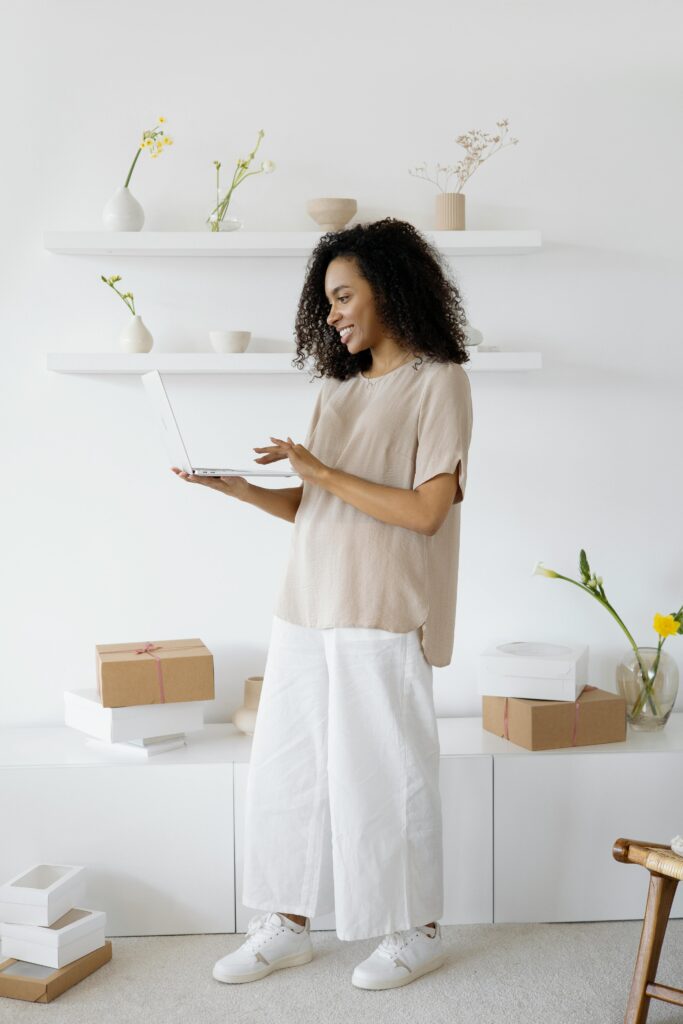 A smiling woman using a laptop surrounded by boxes and decor in a modern, minimalist home office setting.